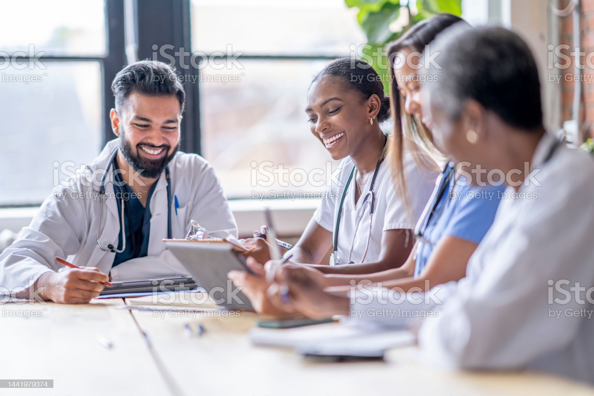 A small group of four medical professionals sit around a boardroom table as they meet to discuss patient cases.  They are each dressed professionally in scrubs and lab coats as they focus on working together.